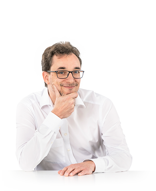Jean-René Thies is standing at a table, his chin resting on one hand. He wears glasses and a white shirt and looks into the camera with a smile.